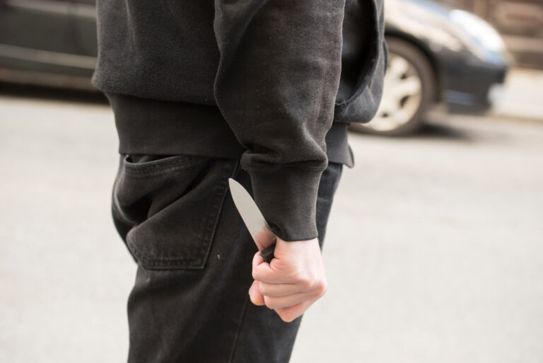 Person concealing knife behind back wearing dark clothing, representing threatening behavior relevant to unlawful assault charges under Victorian law